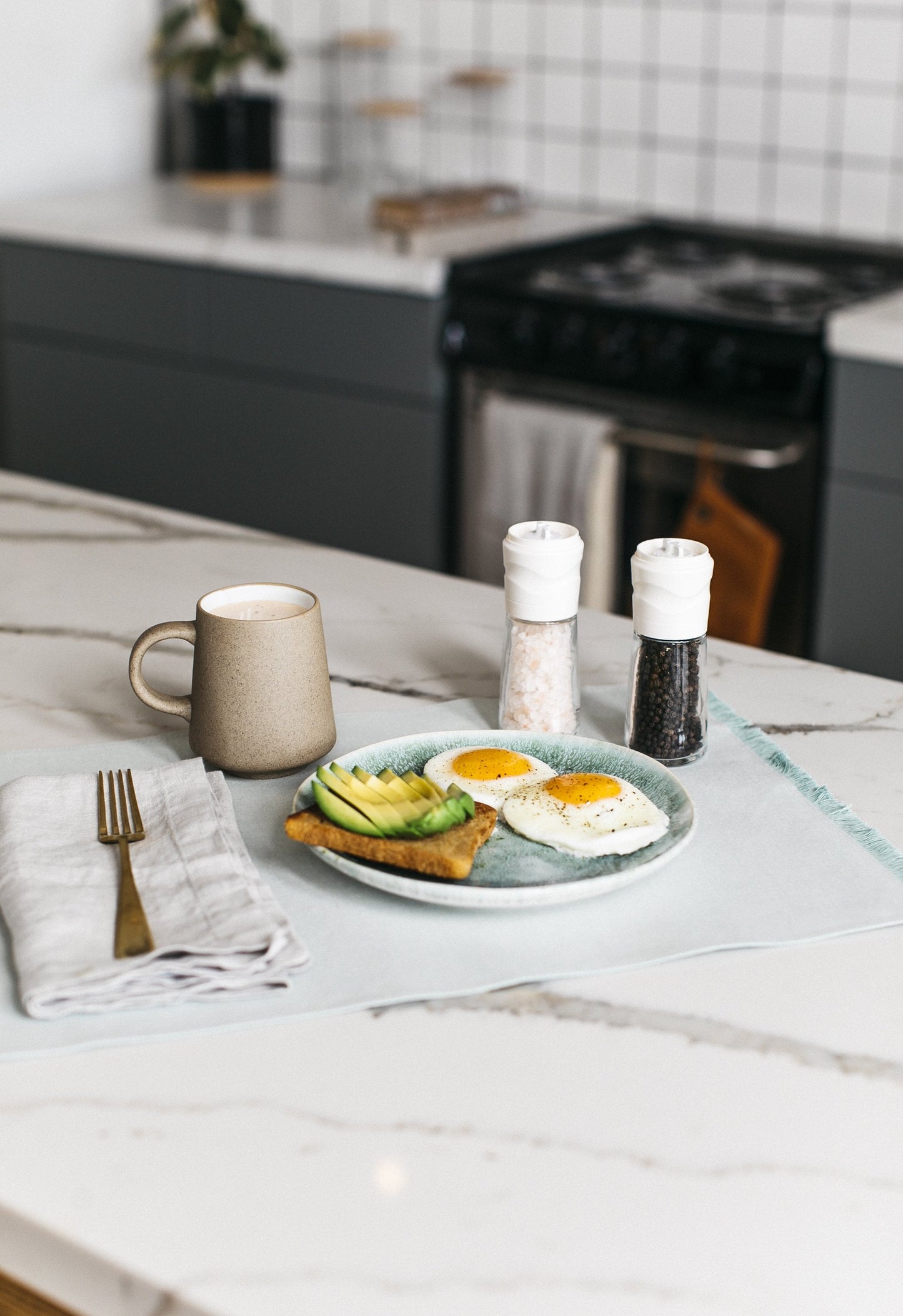 Kyocera Ceramic Salt Grinder right-facing front of Coarse Granulated Culinary Salt product box showing tan mug, on white and black marble kitchen counter with grinder filled with black pepper and grinder filled with salt, gold fork on white cloth napkin, and plate of avocado toast and sunny-side-up eggs