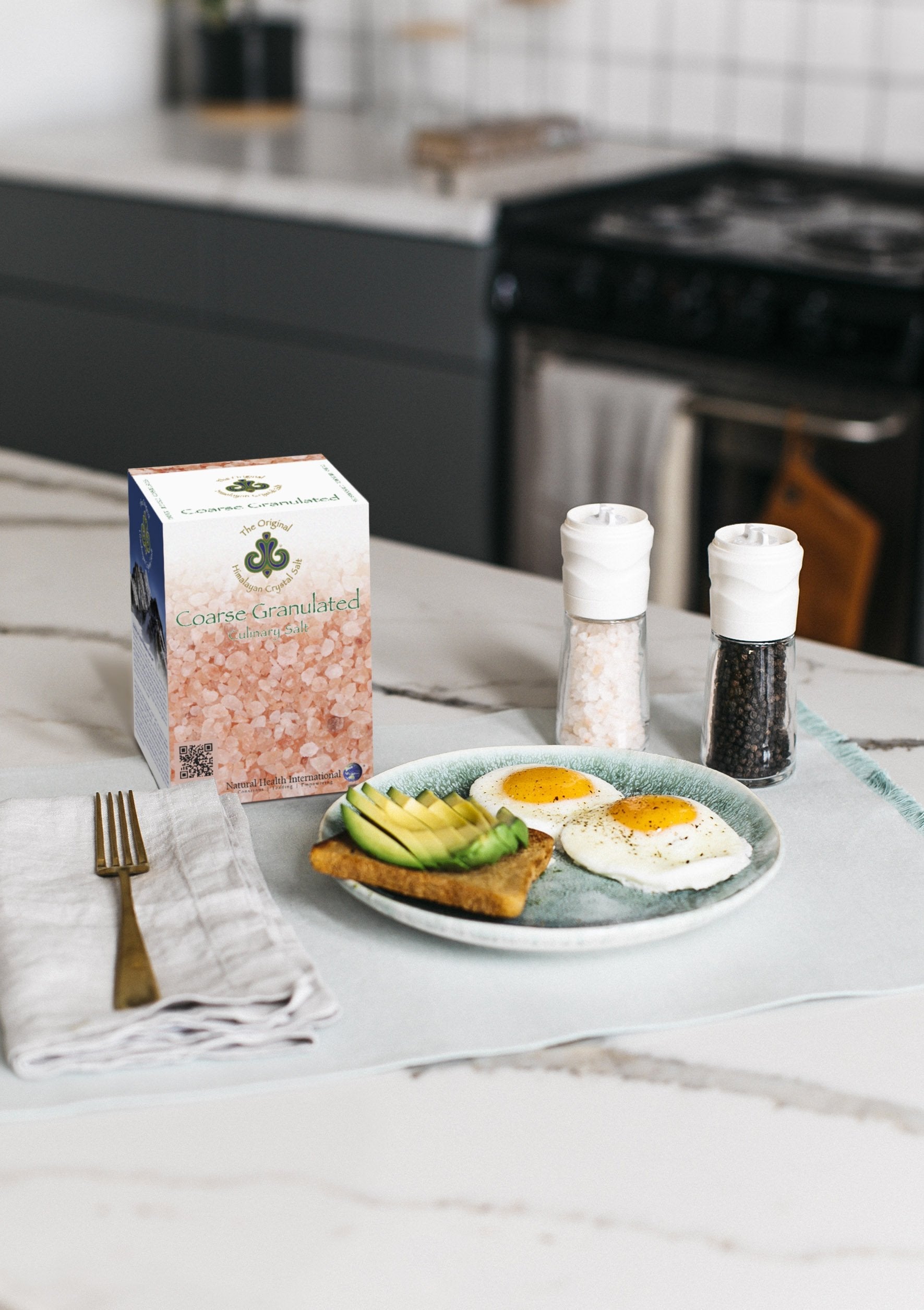 Coarse Salt Bundle (3 Pack) right-facing front of Finely Granulated Culinary Salt product box showing salt granules with salt and spoon, on white and black marble kitchen counter with grinder filled with black pepper and grinder filled with salt, gold fork on white cloth napkin, and plate of avocado toast and sunny-side-up eggs