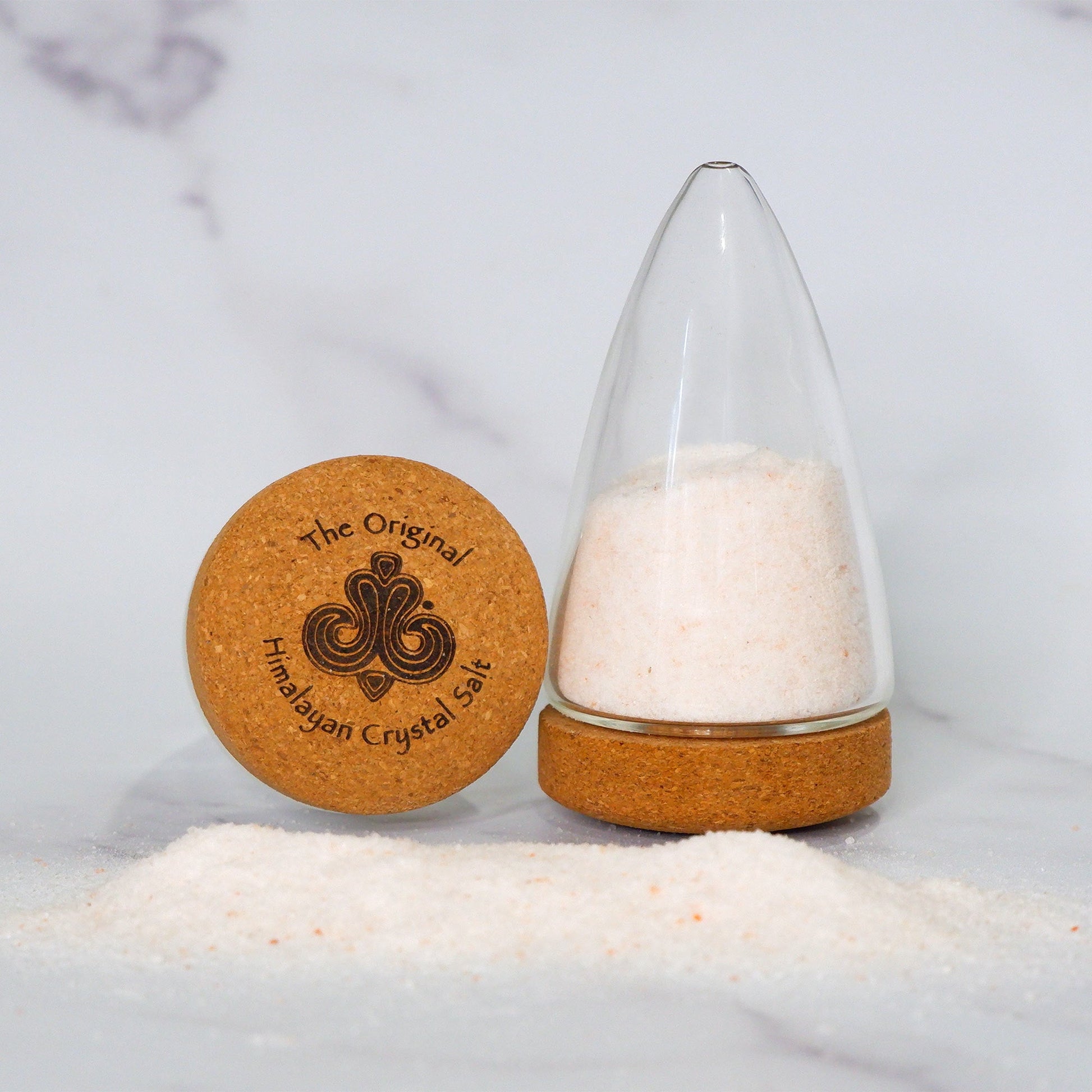 Fine Salt Shaker pointed cylindrical clear glass shaker with cork base with The Original Himalayan Crystal Salt logo, filled with Himalayan crystal salt and more salt on gray countertop