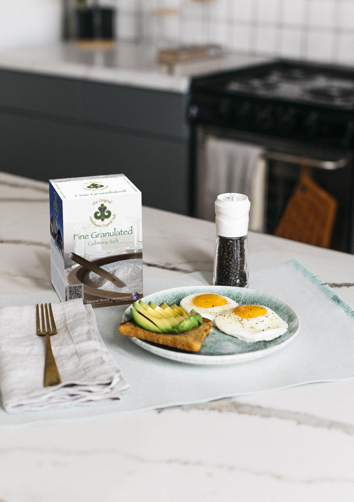 Fine Salt Bundle (3 Pack) right-facing front of Finely Granulated Culinary Salt product box showing salt granules with salt and spoon, on white and black marble kitchen counter with grinder filled with black pepper, gold fork on white cloth napkin, and plate of avocado toast and sunny-side-up eggs