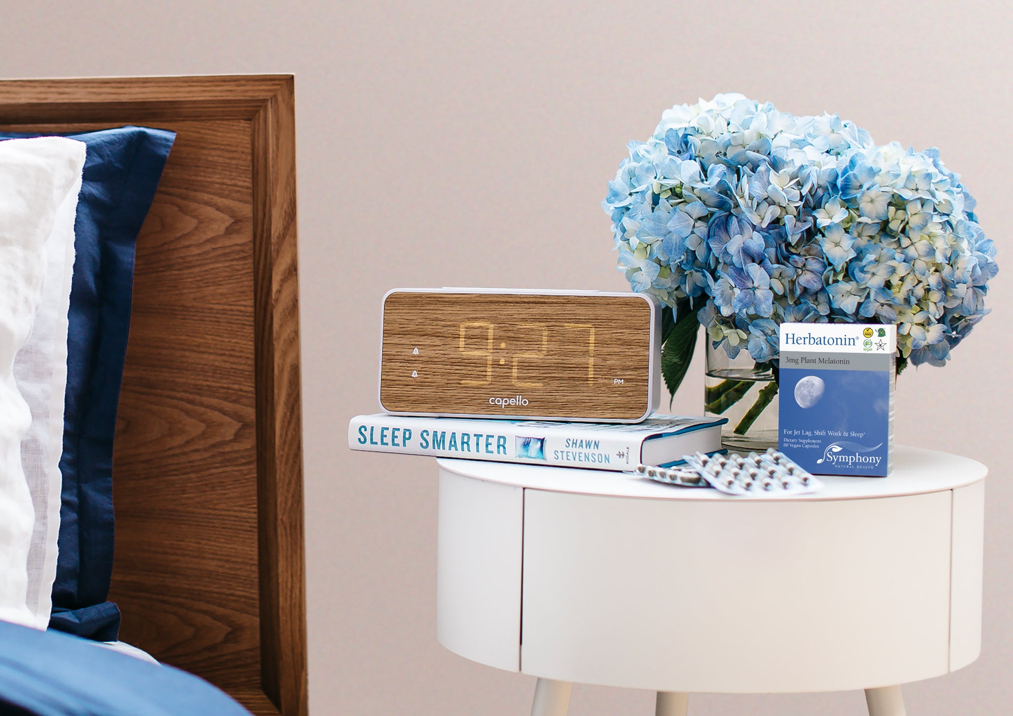 White and Blue bedding with and brown wooden headboard next to white round table with Herbatonin blue box and two blister packs, alarm clock with 9;27 PM digital display on Sleep Smarter by Shawn Stevenson book, blue hydrangea flowers in clear vase filled with water