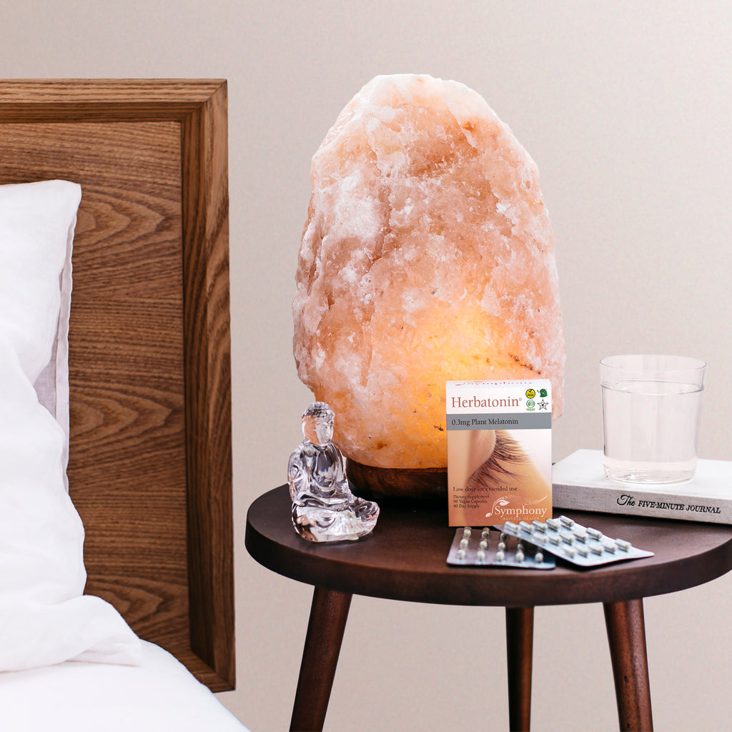 White bedding and brown wooden headboard next to brown wooden table with Herbatonin box and two blister packs, crystal figurine, himalayan crystal salt lamp, glass of water resting on book titled The Five-Minute Journal 