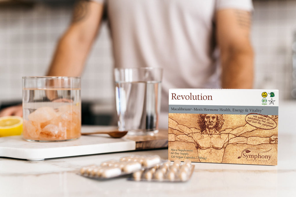 Revolution Macalibrium Man in white top, standing in kitchen with glass sole jar filled with water and sole stones, glass of water, wooden spoon, and Revolution Macalibrium box and blister packs on counter