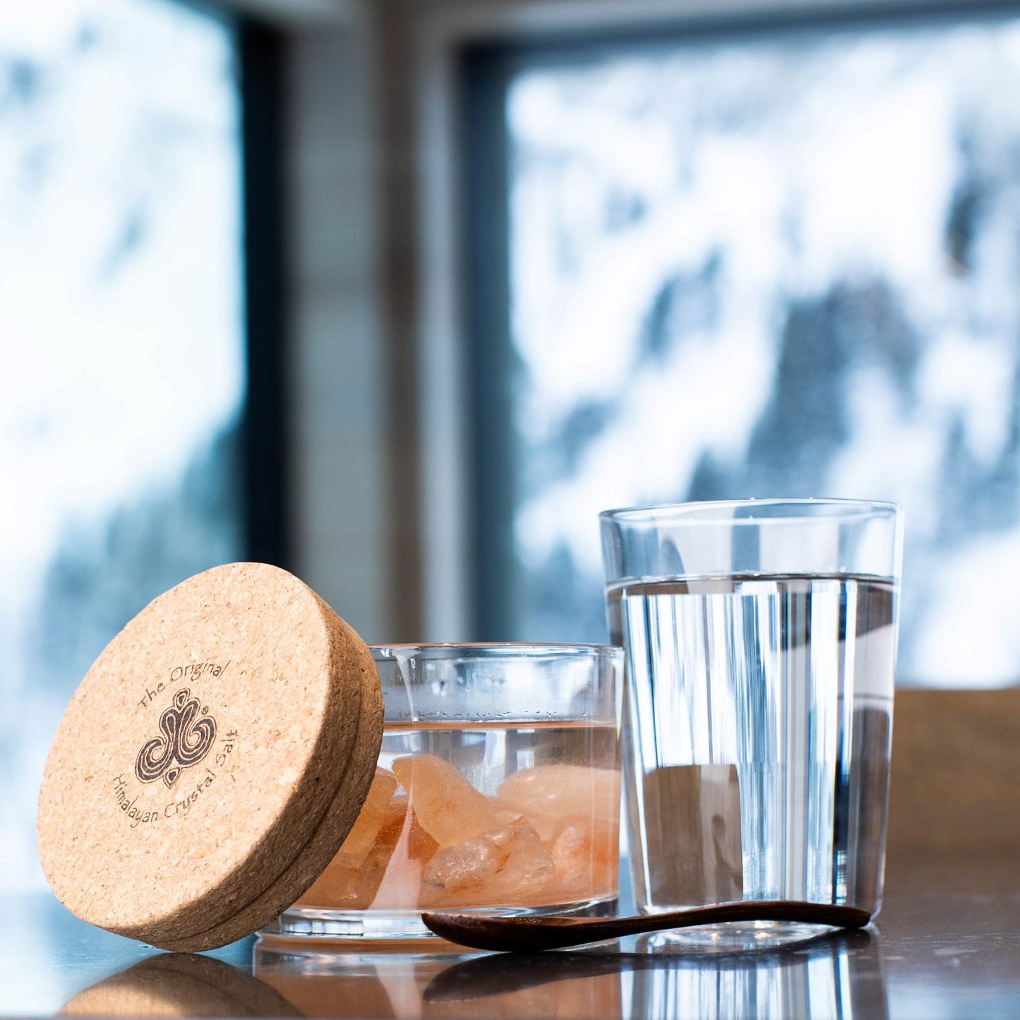 Sole Jar Plus Stones product glass jar filled with Himalayan Crystal Salt stones filled and water, with cork cover, featuring Himalayan Crystal Salt logo glass of water and woodens spoon resting on table with snowy window in background