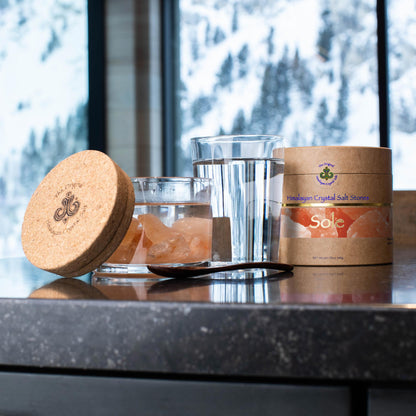 Sole Jar Plus Stones product glass jar filled with water and Himalayan Crystal Salt stones with cork cover and tan product box glass of water and woodens spoon resting on table, both 