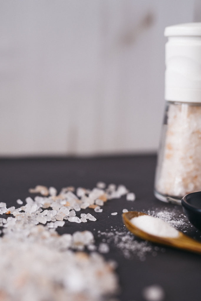 White ceramic and glass salt grinder filled with coarse Himalayan salt, resting on black counter with wooden spoon filled with salt, and salt granules on countertop