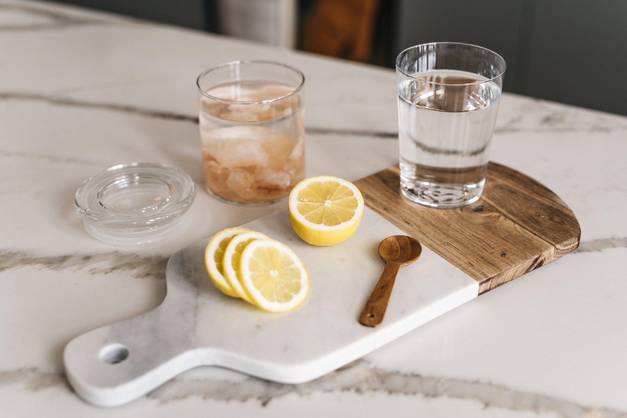 Light gray kitchen counter with glass jar filled with Himalayan Crystal Salt Stones and water, sliced lemons, wooden spoon, glass of water and cutting board