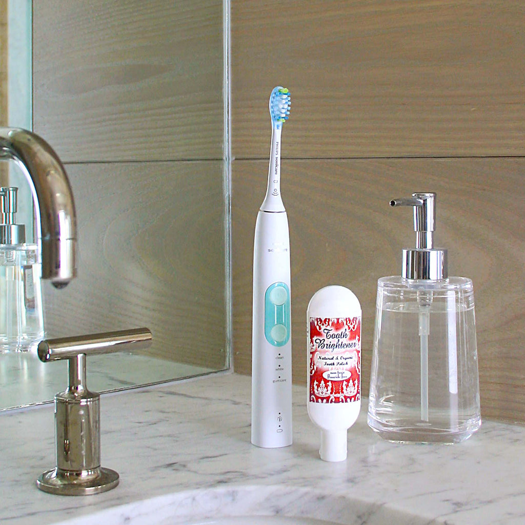 Tooth Brightener Tooth Polish white and turquoise standing electric toothbrush, inverted white bottle with red label, clear glass dispenser, metal faucet, on gray sink ledge, and brown wood wall and mirrored background
