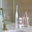 Tooth Brightener Tooth Polish white and turquoise standing electric toothbrush, inverted white bottle with red label, clear glass dispenser, metal faucet, on gray sink ledge, and brown wood wall and mirrored background