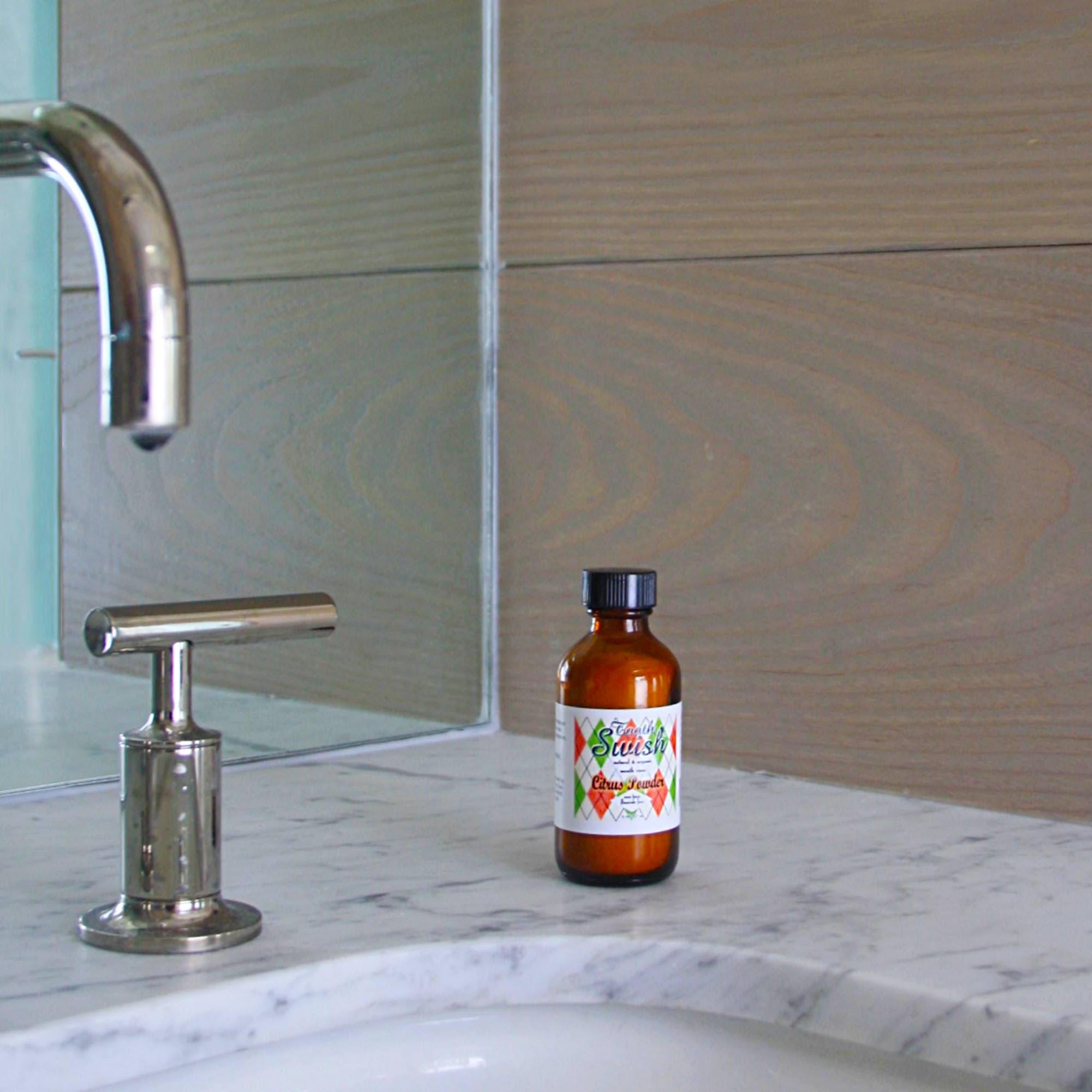 Citrus Tooth Swish brown bottle with black cap with orange, green and white label, resting on sink ledge with metal faucet, on gray sink ledge, and brown wood wall and mirrored background 