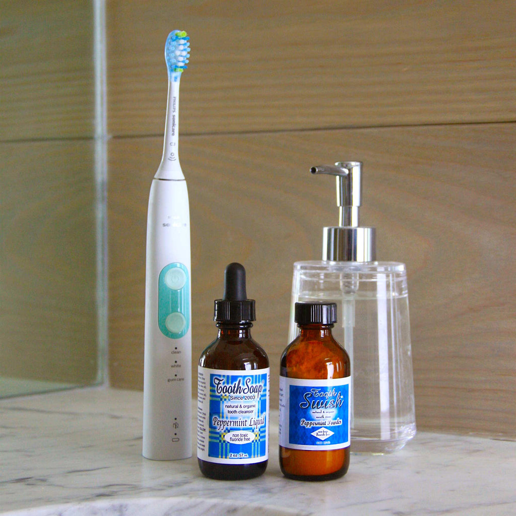 Peppermint Tooth Soap white and turquoise standing electric toothbrush brown bottles with black dropper top, black cap, and blue and white labels, clear glass dispenser