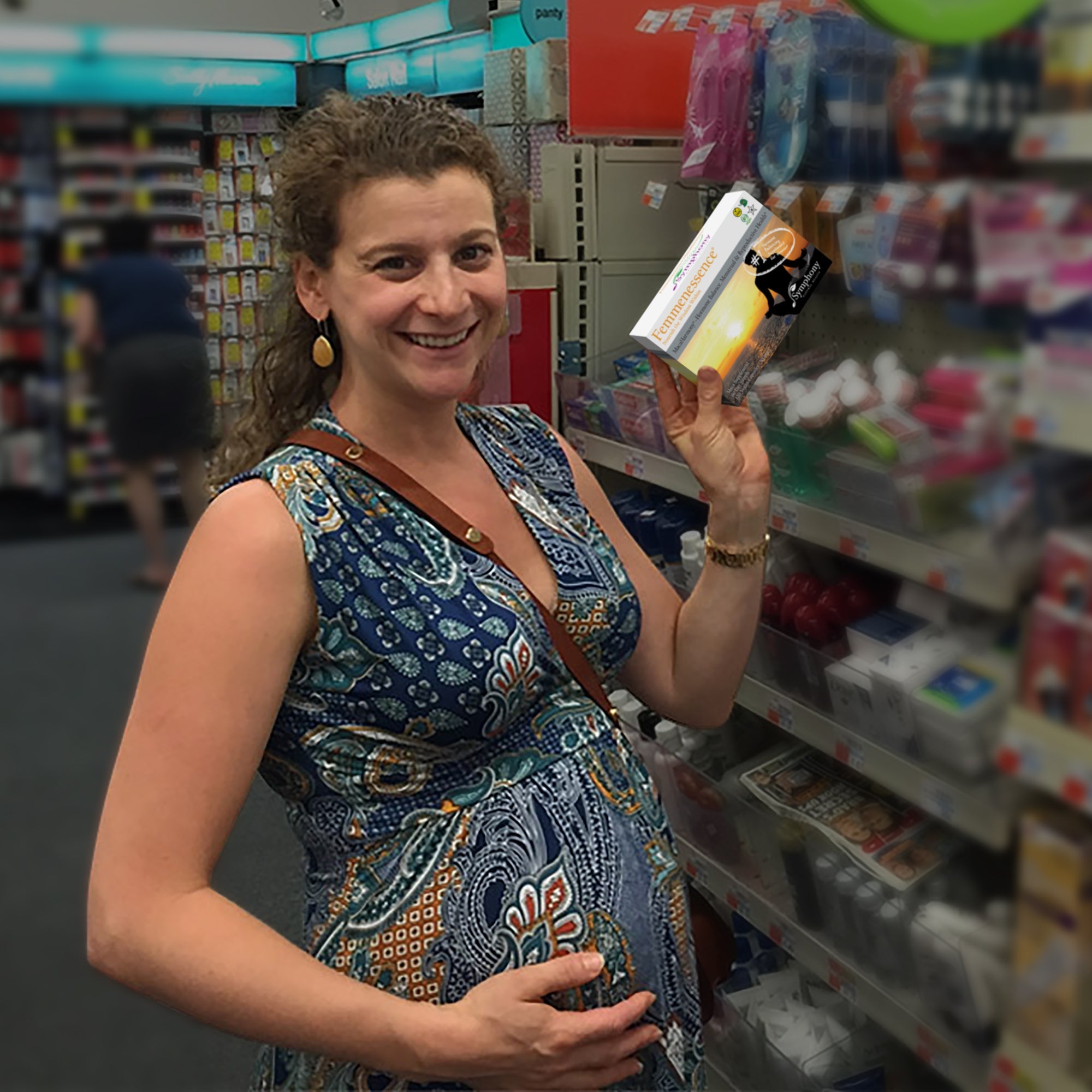 Pregnant white woman in grocery store, with brown curly hair wearing sleeveless blue printed dress, holding stomach and a box of Femmenessence Maca Harmony